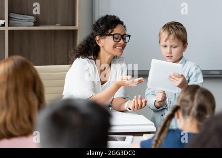Lächelnder afroamerikanischer Lehrer, der in den Händen des Schuljungen auf ein digitales Tablet zeigt Stockfoto