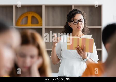 afroamerikanische Lehrerin liest Buch in der Nähe verschwommener Schüler im Klassenzimmer Stockfoto