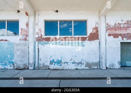 Detailaufnahme einer gebrochenen weißen, blauen und roten Wand eines Hafenlagers mit blauem Himmel, der sich in den langen horizontalen Fenstern widerspiegelt. Altes Gebäude verfällt. Stockfoto