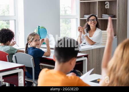 Verschwommenes Schulmädchen, das die Hand in der Nähe der afroamerikanischen Lehrerin und Klassenkameraden hebt Stockfoto
