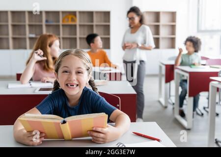 Überglücklich Mädchen hält Buch in der Nähe verschwommene Klassenkameraden und afroamerikanische Lehrerin Stockfoto