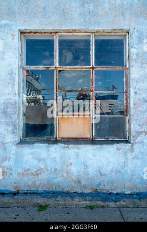 Detailaufnahme eines Fensters eines Hafenlagers mit blauem Himmel und einem Boot, das in seinem quadratischen gefliesten Fenster reflektiert wird. Gesprungene und geschälte weiße, blaue und rote Wand Stockfoto