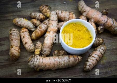 Flache Verlegung von einigen Kurkuma-Wurzeln neben einer Schüssel Kurkuma-Pulver auf einem alten Holzbrett. Dunkles Bild im Food-Stil Stockfoto