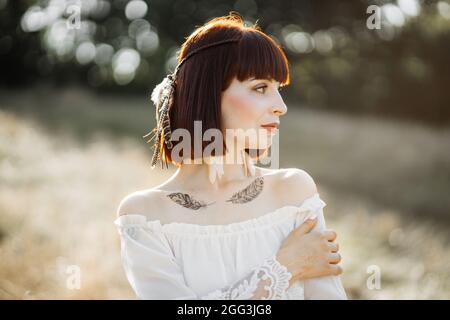 Wilde Natur, Sommer, ethnische Stammesleute. Profil einer verträumten, atemberaubenden jungen Amerikanerin in weißem Kleid, mit Federn in den Haaren und Body Art Tattoos, die auf dem Feld stehen und wegschauen Stockfoto