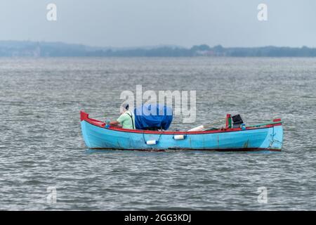 Fischer im grünen T-Shirt, der eine Zigarette raucht, sitzt vor seinem kleinen blauen Boot und segelt auf einem bewölkten Schiff in die Ostsee in der Nähe von Jastarnia, Polen Stockfoto
