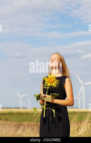 Mädchen mit Sonnenblume in den Händen gegen Feld und Windgeneratoren auf verschwommenem Hintergrund Stockfoto