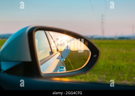 Rückspiegel mit einer Spiegelung der weiblichen Silhouette lädt ein Elektroauto, im Hintergrund sichtbaren Himmel bei Sonnenuntergang Stockfoto