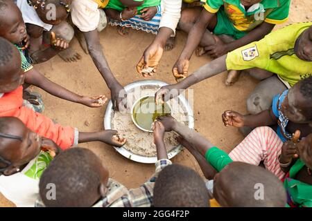 Eine Gruppe von Kindern essen rund um ein gemeinsames Gericht aus Hirsepaste und Baobab-Blatt-Eintopf, wahrscheinlich ihre einzige Mahlzeit des Tages. Diese Großfamilie wurde aufgrund von Gewalt und Unsicherheit vertrieben und steht nun vor Hunger und Nahrungsmittelknappheit. Provinz Kossi, Burkina Faso, Westafrika. Stockfoto