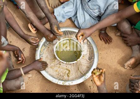 Eine Gruppe von Kindern essen rund um ein gemeinsames Gericht aus Hirsepaste und Baobab-Blatt-Eintopf, wahrscheinlich ihre einzige Mahlzeit des Tages. Diese Großfamilie wurde aufgrund von Gewalt und Unsicherheit vertrieben und steht nun vor Hunger und Nahrungsmittelknappheit. Provinz Kossi, Burkina Faso, Westafrika. Stockfoto