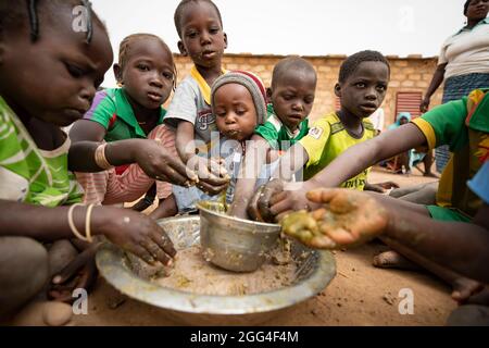 Eine Gruppe von Kindern essen rund um ein gemeinsames Gericht aus Hirsepaste und Baobab-Blatt-Eintopf, wahrscheinlich ihre einzige Mahlzeit des Tages. Diese Großfamilie wurde aufgrund von Gewalt und Unsicherheit vertrieben und steht nun vor Hunger und Nahrungsmittelknappheit. Provinz Kossi, Burkina Faso, Westafrika. Stockfoto