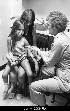 San Antonio, Texas, USA, um 1988: Das junge Mädchen sitzt auf dem Schoß der Mutter und kringt, während es in der städtischen öffentlichen Gesundheitsklinik eine Mumps- und Masernimpfung erhält. ©Bob Daemmrich Stockfoto