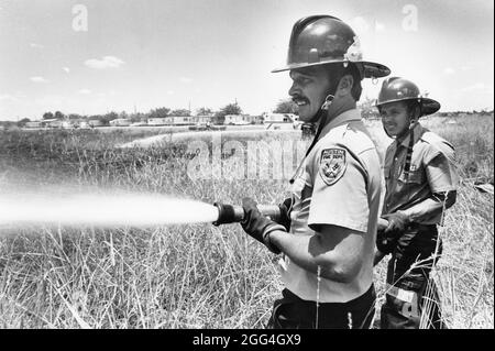 Um 1984 richten die Feuerwehrleute Wasser aus dem Feuerschlauch auf das Grasfeuer in der ländlichen Gegend. ©Bob Daemmrich Stockfoto