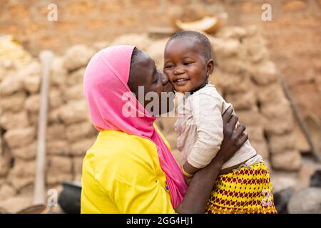 Eine junge Mutter, die einen islamischen Hijab trägt, hebt ihre Tochter auf und küsst sie vor ihrem Haus in Nouna, Burkina Faso, Westafrika. Stockfoto