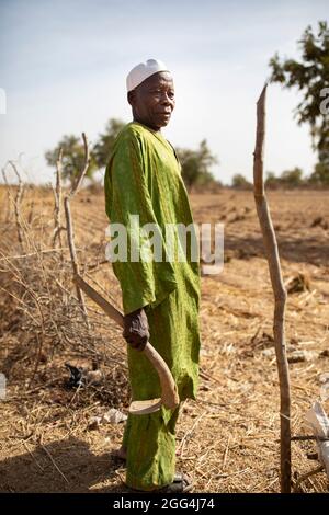 Mahammoud Traore (75) unterstützt eine 21-köpfige Familie durch seine Landwirtschaft im Dorf Dougouninkoro, Mali. Aber der Klimawandel hat die Wettermuster in den letzten Jahren beeinflusst und er konnte nicht so viel wachsen wie früher. Jetzt gehen die Lebensmittelgeschäfte seiner Familie immer aus, bevor er seine neue Ernte ernten kann. Daher gibt es jedes Jahr ein paar Monate, in denen die Familie Hunger hat und sie oft Schulden machen müssen, um Lebensmittel zu kaufen oder sich etwas zu leihen. Hier steht Herr Traore mit einer Hacke in seinen trockenen und kargen Feldern. Sahel-Krise 2021; Barouéli Cercle, Mali. Februar 22, 2021. Foto Stockfoto