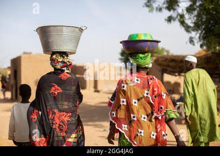 Mahammoud Traore (75) unterstützt eine 21-köpfige Familie durch seine Landwirtschaft im Dorf Dougouninkoro, Mali. Aber der Klimawandel hat die Wettermuster in den letzten Jahren beeinflusst und er konnte nicht so viel wachsen wie früher. Jetzt gehen die Lebensmittelgeschäfte seiner Familie immer aus, bevor er seine neue Ernte ernten kann. Daher gibt es jedes Jahr ein paar Monate, in denen die Familie Hunger hat und sie oft Schulden machen müssen, um Lebensmittel zu kaufen oder sich etwas zu leihen. Hier tragen mehrere Mitglieder von Mr. Traore farbenfrohe Outfits, während sie durch ihr Familiengelände gehen. Sahel-Krise 2021; Barouéli Stockfoto