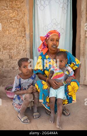 Eine junge alleinerziehende Fulani-Mutter und ihre beiden Töchter sitzen in der Tür ihres Lehmhauses in der Region Ségou, Mali, Westafrika. Stockfoto