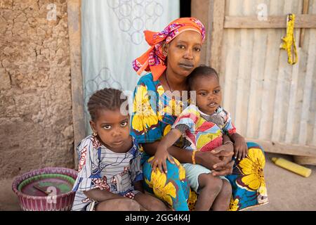 Eine junge alleinerziehende Fulani-Mutter und ihre beiden Töchter sitzen in der Tür ihres Lehmhauses in der Region Ségou, Mali, Westafrika. Stockfoto