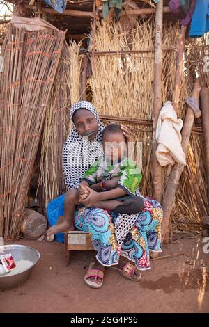 Das Senou Camp liegt am südlichen Rand von Bamako, Malis Hauptstadt. Es ist eines von acht solchen informellen Camps in der ganzen Stadt und allein beherbergt es 223 Familien und etwas mehr als 1.000 Binnenvertriebene. Die hier lebenden Familien sind vor Gewalt und Aufstand im Norden und in den zentralen Teilen des Landes geflohen. Da die meisten von ihnen kein Einkommen oder eine regelmäßige Nahrungsquelle haben, ist der Hunger eine tägliche Herausforderung und die Unterernährung der Bevölkerung ist nach wie vor weit verbreitet. Hier sitzt Fatima Bolly (33) mit ihrem Sohn vor ihrem provisorischen Tierheim, Stockfoto