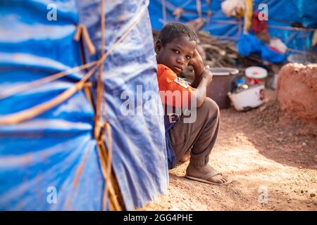 Das Senou Camp liegt am südlichen Rand von Bamako, Malis Hauptstadt. Es ist eines von acht solchen informellen Camps in der ganzen Stadt und allein beherbergt es 223 Familien und etwas mehr als 1.000 Binnenvertriebene. Die hier lebenden Familien sind vor Gewalt und Aufstand im Norden und in den zentralen Teilen des Landes geflohen. Da die meisten von ihnen kein Einkommen oder eine regelmäßige Nahrungsquelle haben, ist der Hunger eine tägliche Herausforderung und die Unterernährung der Bevölkerung ist nach wie vor weit verbreitet. Hier sitzt Moussa Diallo (6) vor dem provisorischen Tierheim seiner Familie. Stockfoto