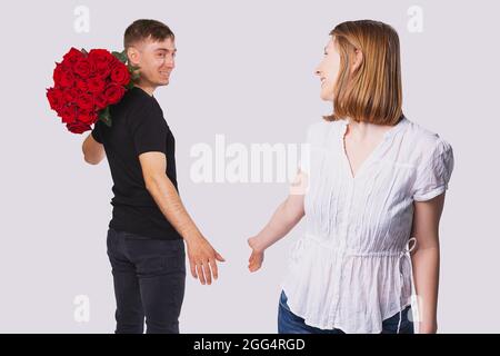 Junges Paar, das in der Ferne steht, Freund, der einen großen Strauß roter Rosen auf der Schulter hält, um es seiner blonden Freundin zu geben. Stockfoto