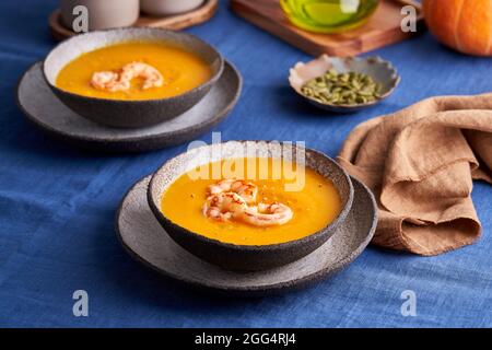 Rustikale hausgemachte Kürbis- und Karotten-Rahm-Püree-Suppe mit Garnelen und Kräutern in einer Schüssel auf der Tischdecke Stockfoto