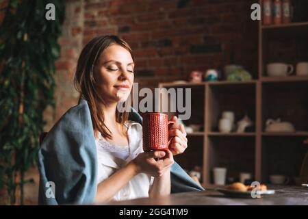 Junge hübsche Frau im gemütlichen grauen Schal mit Tasse Tee in den Händen blickt aus dem Fenster und ruht im Studio Stockfoto