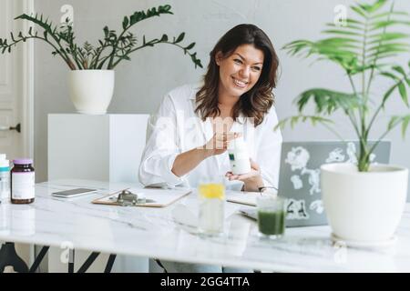 Junge lächelnde Brünette Frau Ernährungsberaterin plus Größe in weißem Hemd arbeiten am Laptop auf dem Tisch mit Haus Pflanze im hellen modernen Büro. Doctor com Stockfoto