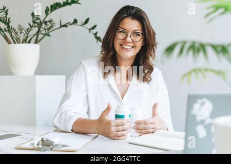 Junge lächelnde Brünette Frau Ernährungsberaterin plus Größe in weißem Hemd arbeiten am Laptop auf dem Tisch mit Haus Pflanze im hellen modernen Büro. Doctor com Stockfoto