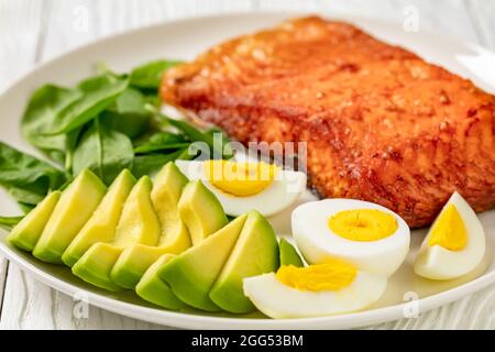 Im Ofen gebackenes Lachsfilet mit cremig reifer Avocado, Babyspinat und hart gekochten Eiern auf einem weißen Teller, gesundes Abendessen Stockfoto