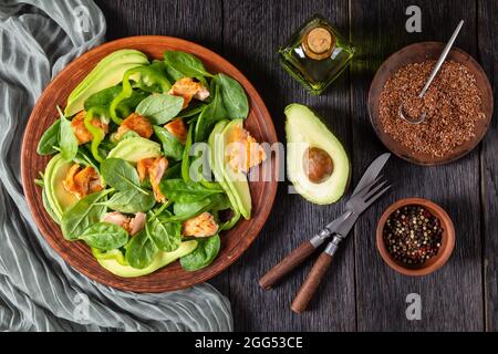 Lachs Spinat Avocado und grünen Pfeffer Salat auf einem braunen rustikalen Teller mit Gabel und Messer auf einem Mooreichentisch mit Zutaten, mediterrane gesunde d Stockfoto