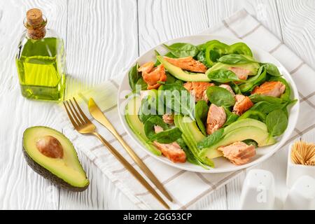 Lachs Spinat Avocado und grünen Pfeffer Salat auf einem weißen Teller mit goldenen Gabel und Messer auf einem weißen Holztisch mit Zutaten, mediterrane Gesundheit Stockfoto
