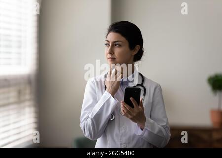 Nachdenkliche junge indische Ärztin, die Smartphone in den Händen hält. Stockfoto