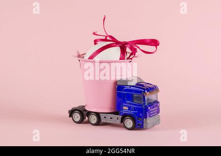 Ein Spielzeug-Traktor transportiert ein Hühnereier in einem Eimer. Blauer Plastikwagen und weißes Ei mit rotem Band. Rosa Hintergrund. Lkw-Transport. Selektiver Fokus. Stockfoto