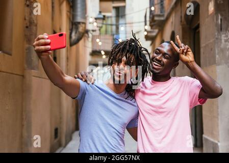 lateinamerikanischer junger Mann mit Dreadlocks, der einen afrikanischen Freund umarmt und sich mit seinem Telefon ein Bild macht Stockfoto