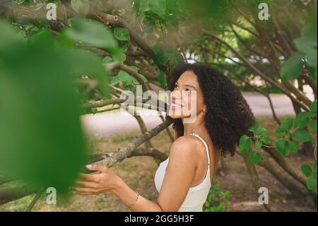 Mode Nahaufnahme Porträt des Lachens Attraktive junge natürlich schöne afroamerikanische Frau mit afrohaaren und perfekten Zähnen Lächeln posiert in Natur Parkland in grünem Laub Stockfoto