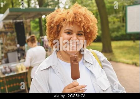 Lifestyle-Porträt von lächelnden jungen natürlich schönen plus size afroamerikanischen Frau mit afro roten Haaren essen Eis Schokolade Popsicle im öffentlichen Park. Vielfalt und körperpositives Konzept Stockfoto