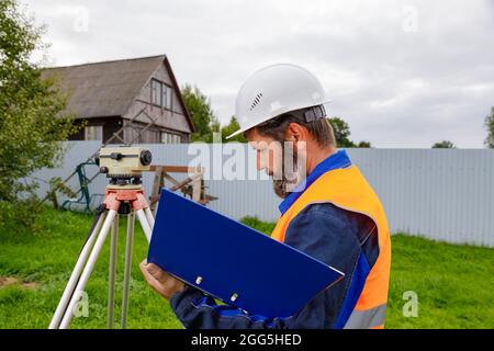 Ein Bauingenieur schreibt die Messwerte der optischen Ebene auf ein Stück Papier in einen Ordner. Ein Mann überprüft die Ebene für den Bau eines Hauses. Stockfoto