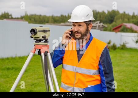 Ein Bauingenieur mit einer optischen Ebene, der auf einem Mobiltelefon spricht. Ein Baumeister in einem Helm mit Bart telefoniert mit Kollegen. Stockfoto