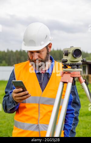 Ein Bauingenieur mit einer optischen Ebene schaut in ein Mobiltelefon. Ein bärtiger Mann sucht auf seinem Handy nach Informationen. Stockfoto
