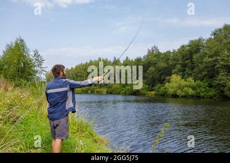 Ein Mann mit Bart fischt auf dem Fluss und wirft eine Leine. Ein Fischer mit einer Angelrute fischt am Flussufer. Stockfoto