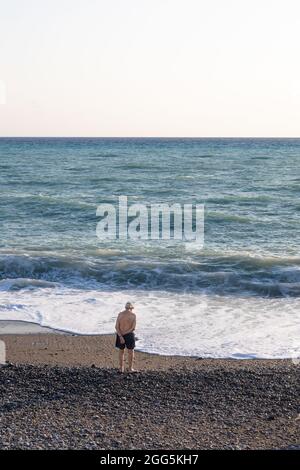 Der alte Mann steht allein vor dem Meer, die Hände von hinten gekreuzt Stockfoto