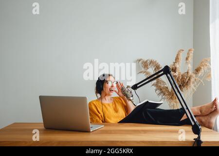 Die junge Radiomoderatorin sitzt mit den Füßen auf seinem Schreibtisch und nimmt Audio-Podcasts auf. Weibliche Podcasterin streamt ihre Stimme zu Hause ins Mikrofon Stockfoto