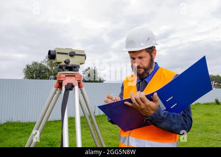 Ein Bauingenieur schreibt die Messwerte der optischen Ebene auf ein Stück Papier in einen Ordner. Ein Mann überprüft die Ebene für den Bau eines Hauses. Stockfoto