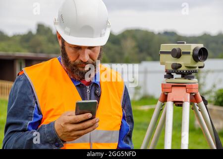 Ein Bauingenieur mit einer optischen Ebene schaut in ein Mobiltelefon. Ein bärtiger Mann sucht auf seinem Handy nach Informationen. Stockfoto