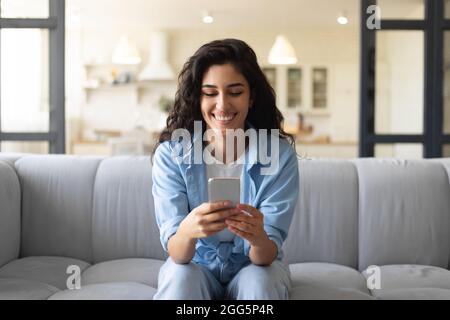 Glückliche junge Frau, die mit dem Smartphone auf der Couch sitzt, soziale Medien überprüft, im Internet surft, von zu Hause aus online chattet Stockfoto