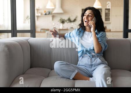 Fröhliche junge Brünette Dame auf dem Sofa sitzen, Anruf auf Handy zu Hause, kopieren Raum Stockfoto
