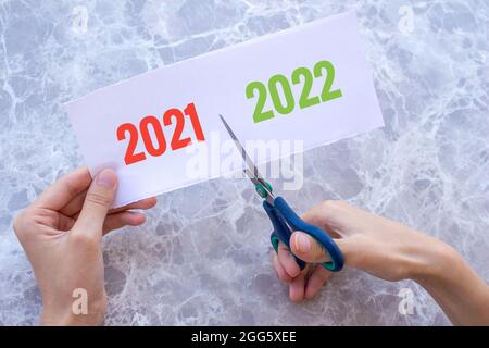 Die Hand einer Frau schneidet das Papier mit den Worten 2021 und 2022 mit einer Schere. Neues Jahr 2022 kommt Konzept. Neues Jahr Neubeginn. Stockfoto