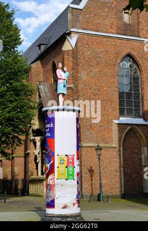 Lebensechte Säulenheilige Skulptur einer Mutter mit ihrem Kind vom Künstler Christoph Pöggeler. St. Lambertus Kirche im Hintergrund. Stockfoto