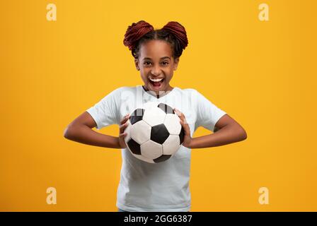 Fröhliches afro-amerikanisches Mädchen, das den Fußball auf Gelb hält Stockfoto