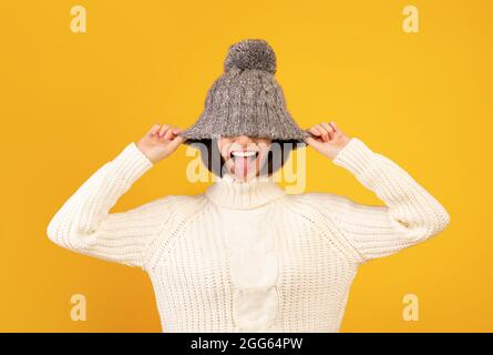 Verspielte tausendjährige Frau, die einen Wollhut herunterzieht und ihre Zunge herausstreckt und über gelben Hintergrund duckt Stockfoto